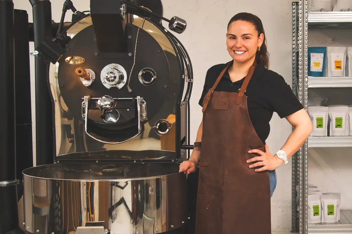 Kvinne i brunt forkle smiler ved siden av en stor kaffebrennemaskin for bedrifter i industrielle omgivelser.