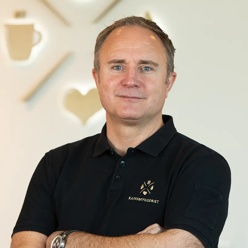 En mann i svart poloskjorte står med armene i kors og smiler lett. Skjorten viser logo og tekst til Organisasjon Kaffebryggeriet. Bakgrunnen kompletterer scenen med innredning med kaffetema.