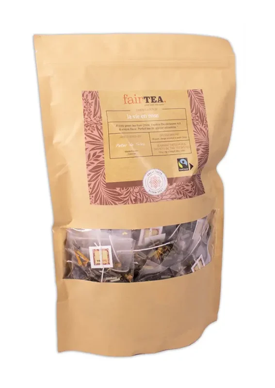 Gylden papirpakke med FairTea la vie en rose-te, med et klart vindu som avslører teposer inni, utsmykket med etiketter og en fair trade-logo.