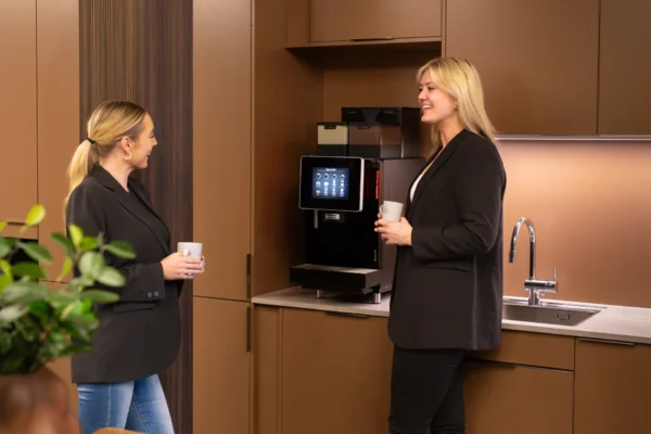 To kvinner i businessantrekk står i et moderne kjøkken med krus i hendene og diskuterer riktig kaffe og kaffemaskin til kontoret ved kaffemaskinen på kjøkkenbenken.