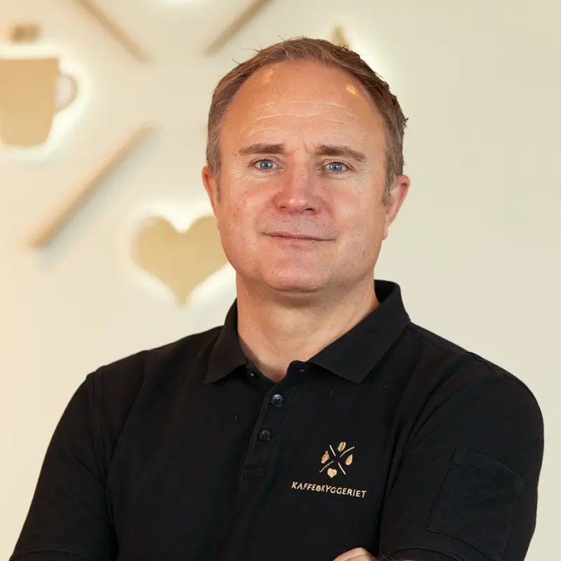 En person i svart poloskjorte med emblem fra Organisasjon Kaffebryggeriet står med armene i kors mot en lys bakgrunn med innredning i kaffetema.