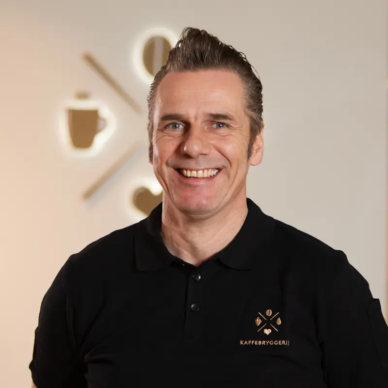 En middelaldrende mann med et vennlig smil iført svart poloskjorte brodert med Organisasjon Kaffebryggeriets logo, stående foran en beige bakgrunn med klokkedesign.