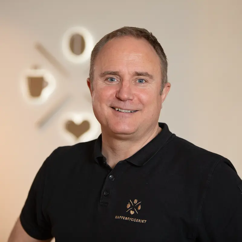 Middelaldrende mann med kort hår, smilende, iført svart poloskjorte med Organisasjon Kaffebryggeriets logo, stående i et rom med dekorativ veggklokke og hjertesymbol i