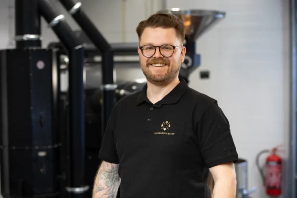 En mann med briller og tatoveringer, iført svart polo med kaffebrenneri-logo, står smilende i et industrielt miljø og tilbyr stolt kaffeløsning til bedrift blant kaffebrenneriutstyr i bakgrunnen.