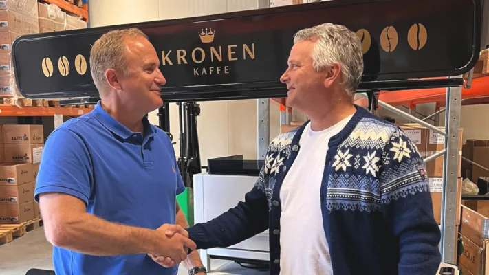 To menn håndhilser foran en kaffemaskin med påskriften «KRONEN KAFFE» inne i det som ser ut til å være et lager eller et industrilokale, tydelig fornøyde med valget av riktig kaffemaskin til kontoret.