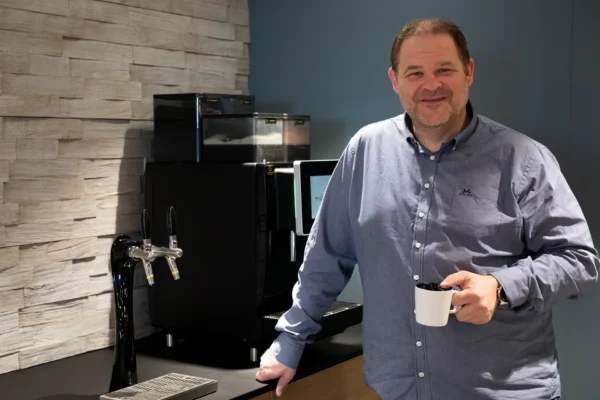 En mann i blå skjorte står ved en kaffemaskin, holder et hvitt krus og smiler, og viser frem en kaffeløsning for bedrift i et moderne kjøkken- eller pauseromsmiljø.