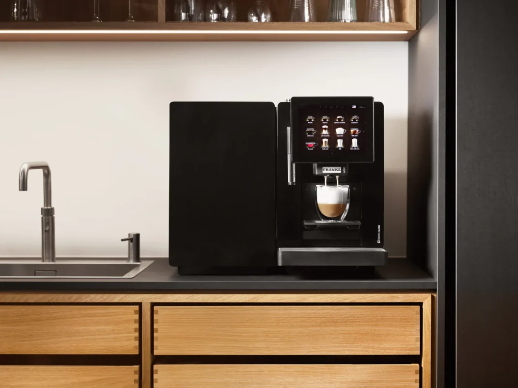 En ny kaffemaskin i elegant svart tilbereder en kopp kaffe på kjøkkenbenken ved siden av en vask og skuffer i tre, med hyller med glassvarer over.