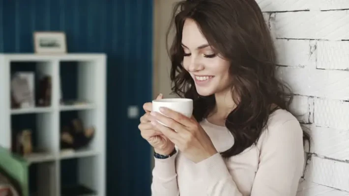 Kvinne smiler forsiktig mens hun holder en Bravilor Bonamat Esprecious 22 kaffemaskin hvit kaffekopp, stående mot en hvit murvegg med blå bakgrunn.
