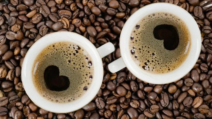 To hvite kaffekopper fylt med svart kaffe fra en ny kaffemaskin står på en seng av ristede bønner, og hver kopp har et hjerteformet mønster i cremaen.