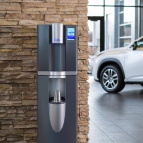 En moderne vanndispenser står mot en steinsøyle i et bilforhandlerutstillingsrom med kjøretøy synlige i bakgrunnen.