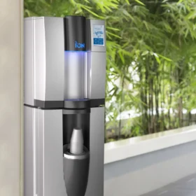 Moderne ION SODA vanndispenser med digital temperaturvisning og fyllområdebelysning, plassert i en bygningslobby med frodige grønne planter i bakgrunnen.