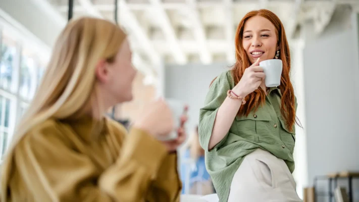 To kvinner sitter innendørs med kaffekrus fra en kaffemaskin øker produktiviteten i hånden og snakker sammen i et lyst rom med store vinduer.
