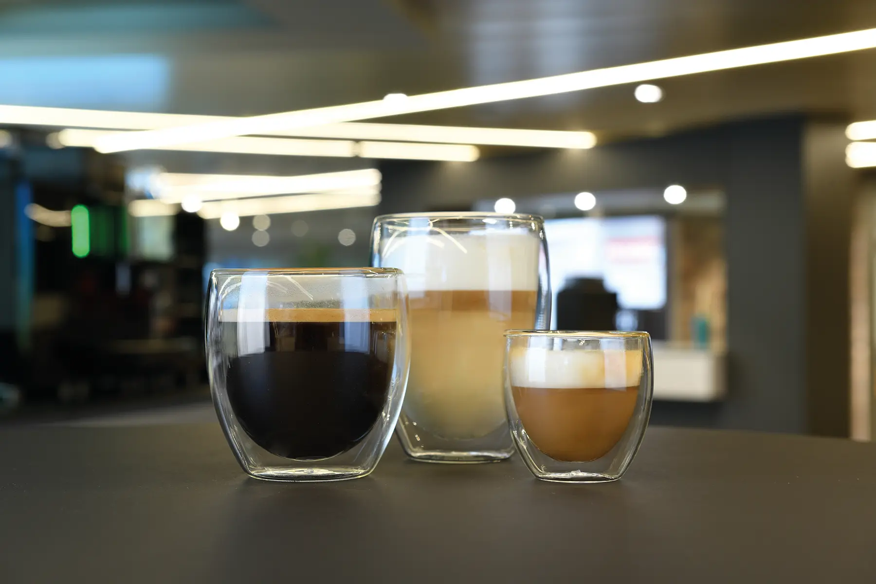 Tre dobbeltveggede glasskopper – svart kaffe, latte og cappuccino – sitter på en mørk overflate i en moderne innendørs setting, perfekt for de som lurer på "hva koster kaffemaskin" for å nyte kafédrinker hjemme.