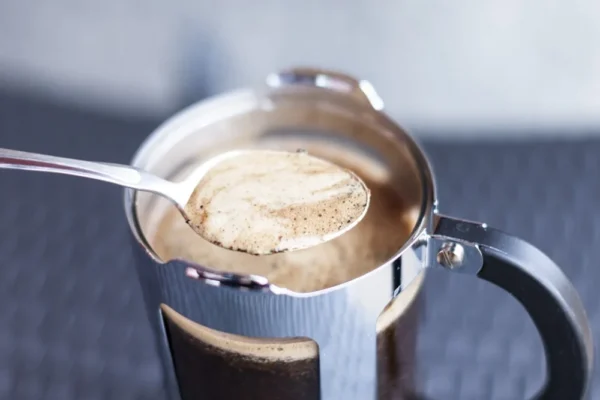 En skje som løfter skummende kaffe fra en presskannekaffe i rustfritt stål.