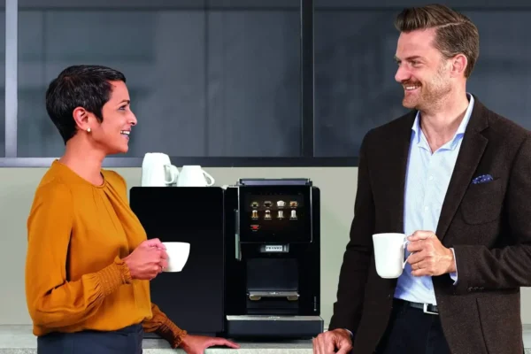 To fagfolk, en mann og en kvinne, smiler og snakker mens de holder kaffekopper ved siden av en kaffemaskin på kontoret, og diskuterer hvorfor investere i en kaffemaskin.