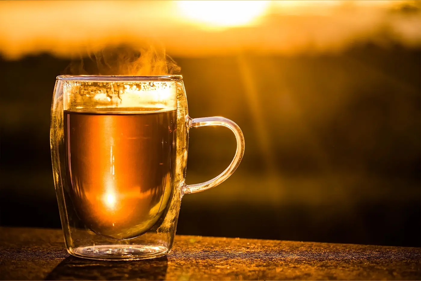 Et glasskrus med varm te med damp som stiger, plassert på en treoverflate på kontoret mot en varm solnedgangsbakgrunn.