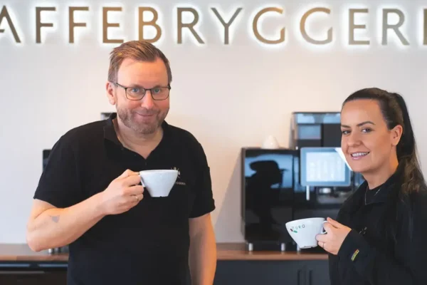 To smilende baristaer holder kaffekopper på en kafé med ordet "kaffebryggeriet" opplyst på veggen bak, stående ved siden av en kaffemaskin.