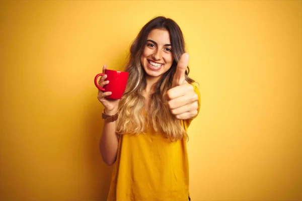 En smilende kvinne i gul skjorte holder en rød kopp kaffe og gir tommel opp mot en gul bakgrunn.