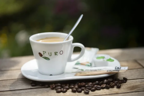 En kopp espresso med tallerken og skje, bærekraftige puro-kaffepakker på siden, omgitt av kaffebønner på et trebord.