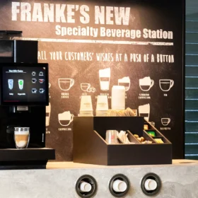En moderne kaffemaskin og krydderstasjon, merket "FRANKE'S NEW Specialty Beverage Station", med Franke SB1200 kaffemaskin, er satt opp på en benkeplate. De tilbyr ulike kaffe- og tealternativer med en illustrert guide bak seg.