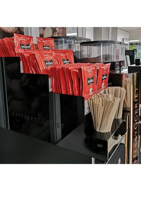 En rekke røde miko-kaffepakker ved siden av trerørere og kaffedispensere på en selvbetjent stasjon.