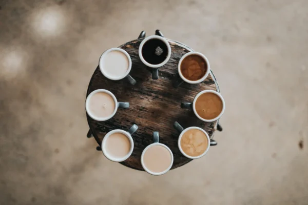Toppvisning av et rundt trebord med åtte forskjellige kopper kaffe, som danner en sirkel, på et betonggulv.